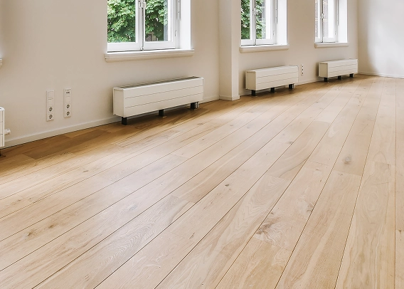 Bright, empty room with light wooden flooring by State Floors, white walls, three large windows, and three white radiators under the windows. Green foliage is visible outside, bringing a touch of Rhode Island nature indoors.
