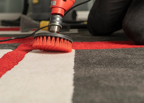 A person uses an electric scrubbing brush with orange bristles to clean a red, white, and gray carpet from State Floors, focusing on a stained area.