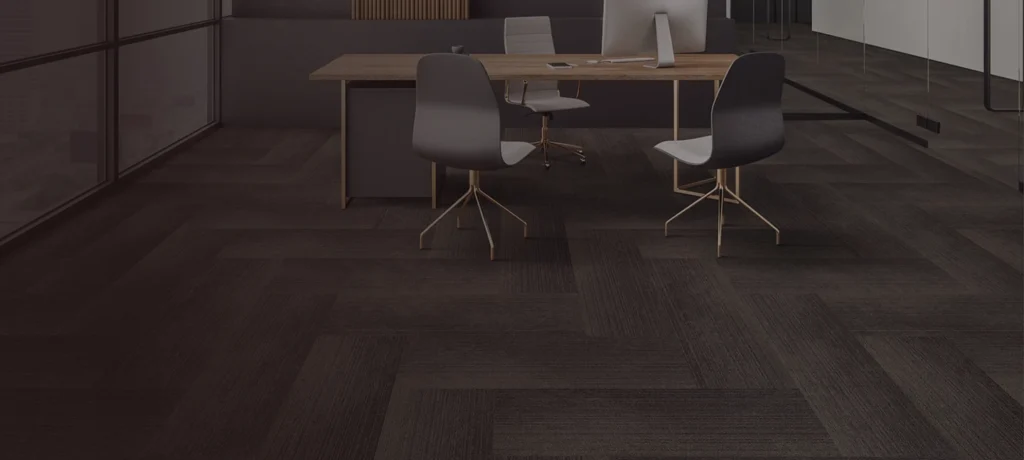 A modern office with a wooden desk, two gray chairs on wheels, an all-in-one desktop computer, and dark herringbone-patterned State Floors carpeting. Large windows and sleek partitions line the Rhode Island workspace.